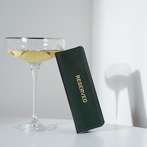 May include: A green leather table card with gold lettering that reads "RESERVED" and a champagne flute filled with a pale yellow liquid.