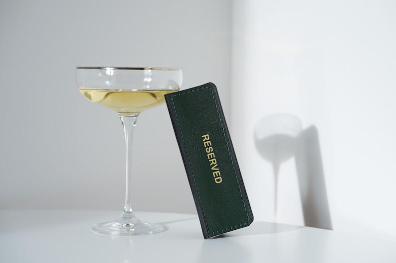 May include: A green leather table card with gold lettering that reads "RESERVED" and a champagne flute filled with a pale yellow liquid.
