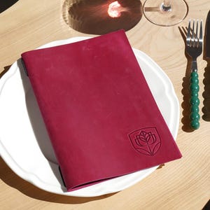 May include: A deep magenta leather menu cover featuring an embossed shield logo with a stylized flower design. The rectangular cover rests on a white scalloped plate next to a silver fork and knife with green beaded handles on a light wood surface.
