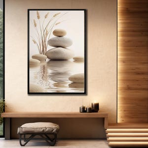 Op de afbeelding: Een ingelijste kunstdruk met een serene scène van gestapelde stenen en tarwearen die weerspiegeld worden in het water. Het kunstwerk heeft een neutraal kleurenpalet met beige, grijze en bruine tinten, wat een rustgevende en minimalistische esthetiek creëert.