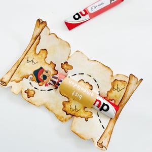 Puede incluir: Una barra de caramelo roja y blanca envuelta en un rollo de papel marr&oacute;n con el texto "Bist eingeladen" en alem&aacute;n. El rollo tiene forma de mapa del tesoro con una ilustraci&oacute;n de pirata.