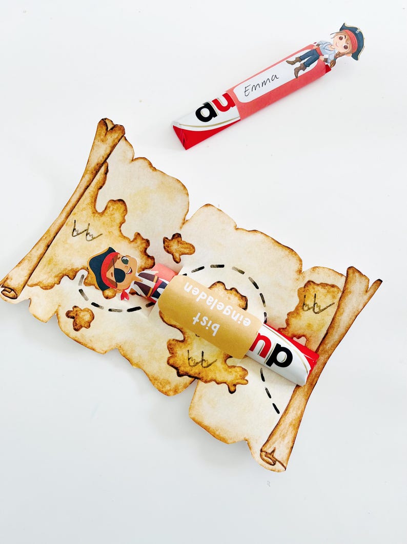 Puede incluir: Una barra de caramelo roja y blanca envuelta en un rollo de papel marr&oacute;n con el texto "Bist eingeladen" en alem&aacute;n. El rollo tiene forma de mapa del tesoro con una ilustraci&oacute;n de pirata.
