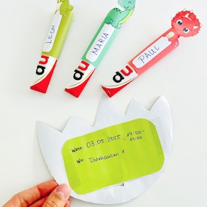 May include: Three colorful dinosaur-themed party favors with names "LEON", "MARIA", and "PAUL" on them. Each favor has a different dinosaur design and is attached to a red and white wrapper. A green invitation with text is held in the hand.