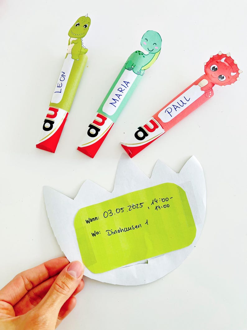 May include: Three colorful dinosaur-themed party favors with names "LEON", "MARIA", and "PAUL" on them. Each favor has a different dinosaur design and is attached to a red and white wrapper. A green invitation with text is held in the hand.