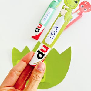 May include: A hand holding a green and white dinosaur-themed bookmark with the name "LEON" on it. Two other dinosaur bookmarks are visible, one with the name "MARIA" and another red one. The bookmarks are on a white background.