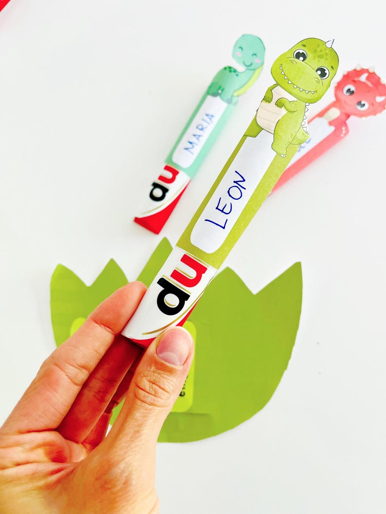 May include: A hand holding a green and white dinosaur-themed bookmark with the name "LEON" on it. Two other dinosaur bookmarks are visible, one with the name "MARIA" and another red one. The bookmarks are on a white background.