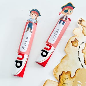 Puede incluir: Dos barras de caramelo rojas y blancas con envoltorios de papel rosa. Los envoltorios tienen los nombres "Emma" y "Lukas" escritos en ellos. Cada barra de caramelo tiene un personaje de pirata de dibujos animados en la parte superior.
