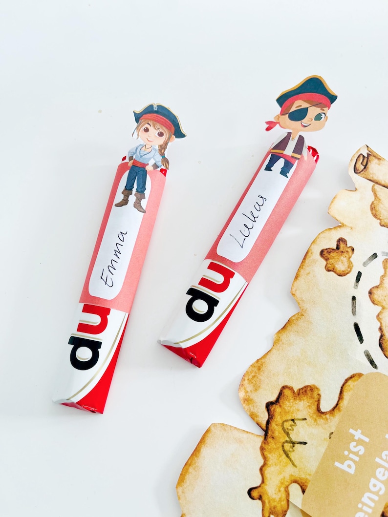 Puede incluir: Dos barras de caramelo rojas y blancas con envoltorios de papel rosa. Los envoltorios tienen los nombres "Emma" y "Lukas" escritos en ellos. Cada barra de caramelo tiene un personaje de pirata de dibujos animados en la parte superior.