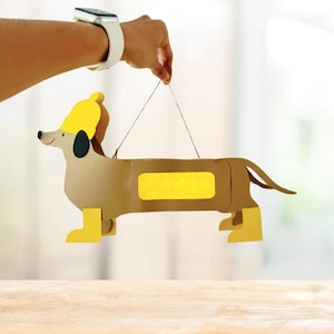 Könnte beinhalten: Eine handgefertigte Dackel-Laterne. Die Laterne ist hauptsächlich braun mit gelben Akzenten auf Hut, Stiefeln und rechteckigem Fenster. Der Hund hat ein schwarzes Ohr und ein lächelndes Gesicht. Sie wird an einer Schnur aufgehängt.