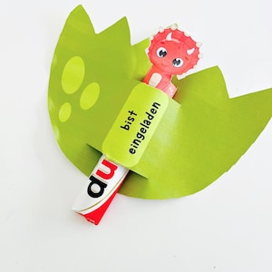 May include: A green paper dinosaur egg with a red and white candy bar and a cartoon dinosaur. The candy bar has the word "du" on it. The green paper has the words "bist eingeladen" on it.