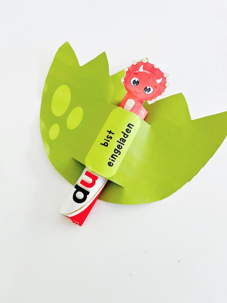 May include: A green paper dinosaur egg with a red and white candy bar and a cartoon dinosaur. The candy bar has the word "du" on it. The green paper has the words "bist eingeladen" on it.