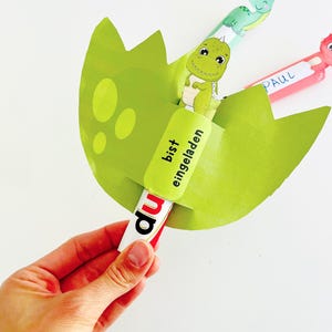 May include: A green dinosaur-themed invitation card shaped like a cracked egg, held by a hand. The card features a cartoon dinosaur and the German text "bist eingeladen" and "du". Additional cards with dinosaur illustrations are visible.