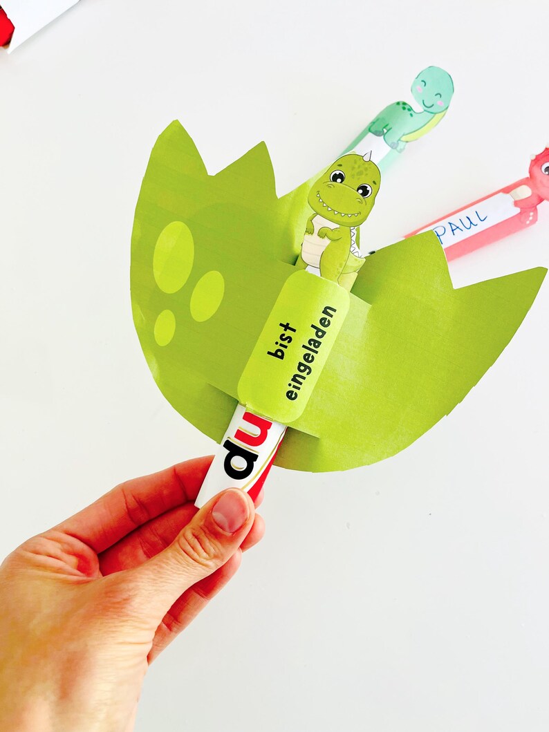 May include: A green dinosaur-themed invitation card shaped like a cracked egg, held by a hand. The card features a cartoon dinosaur and the German text "bist eingeladen" and "du". Additional cards with dinosaur illustrations are visible.