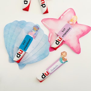 Könnte beinhalten: Duplo-Schokoriegel mit Dekorationen zum Thema Meerjungfrau. Die Dekorationen umfassen eine blaue Muschel, einen rosa Seestern und einen Schokoriegel mit dem Namen "Isabelle". Die Riegel tragen den Text "du" und "bist eingeladen".