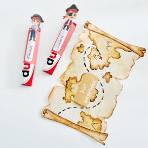 Puede incluir: Dos barras de caramelo rosa y rojo con personajes de piratas en la parte superior. Las barras de caramelo est&aacute;n etiquetadas con "du" y tienen los nombres "Emma" y "Lucas" escritos en ellas. Un mapa del tesoro marr&oacute;n y blanco con una X roja y el texto "bist eingeladen" est&aacute; en el fondo.