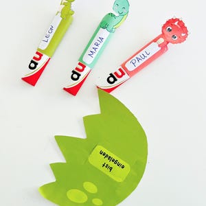 May include: Three colorful dinosaur-themed party favors with the names Leon, Maria, and Paul. Each favor has a cartoon dinosaur and the word "du" printed on it. A green, egg-shaped cutout with the text "Bist eingeladen" is also present.