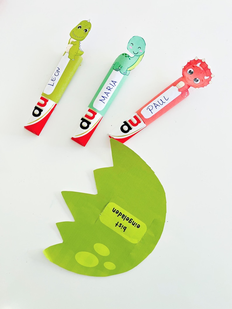 May include: Three colorful dinosaur-themed party favors with the names Leon, Maria, and Paul. Each favor has a cartoon dinosaur and the word "du" printed on it. A green, egg-shaped cutout with the text "Bist eingeladen" is also present.