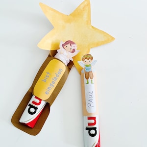 以下が含まれることがあります： 妖精のイラストと「Paul」の文字が書かれた2本のチョコレートバー。チョコレートバーは茶色の紙ホルダーに入っており、背景には金の星があります。1本のチョコレートバーには「bist eingeladen」という文字が書かれています。