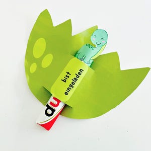 May include: A green paper dinosaur hatching from a green egg, with the words "bist eingeladen" on a yellow label. A red and white candy bar is partially visible.