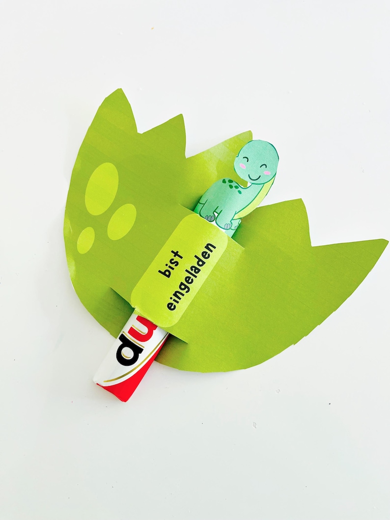 May include: A green paper dinosaur hatching from a green egg, with the words "bist eingeladen" on a yellow label. A red and white candy bar is partially visible.