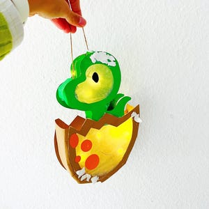 Könnte beinhalten: Ein grüner Dinosaurier schlüpft aus einem gelben Ei mit roten Flecken. Der Dinosaurier hat ein Licht im Inneren und hängt an einer Schnur.