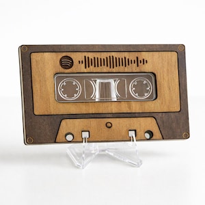 May include: A wooden cassette tape design with a Spotify logo and soundwave graphic. The cassette is two-toned, with a lighter wood grain center and a darker wood grain border. It is displayed on a clear stand.