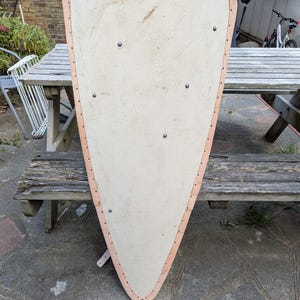 May include: A wooden shield with a light tan surface and a light brown leather trim. The shield is roughly triangular with a rounded top and is secured with rivets. A leather strap is visible on the back.