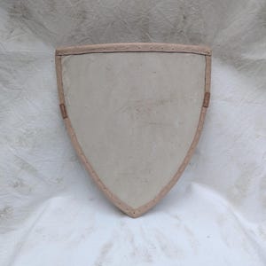 May include: A shield-shaped item with a light tan interior and a darker tan border. The border is stitched with a contrasting thread. Two small, rectangular tan leather pieces are attached to the shield.