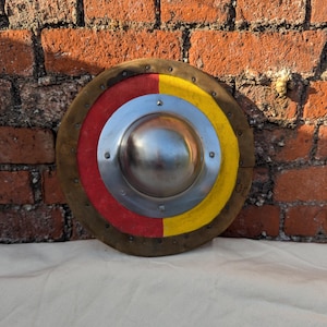 Pode incluir: Um escudo redondo com um chefe central de metal e uma estrutura de madeira. O escudo é pintado com segmentos vermelhos e amarelos e tem uma borda de couro castanho. O escudo é provavelmente para reconstituição histórica ou exibição.