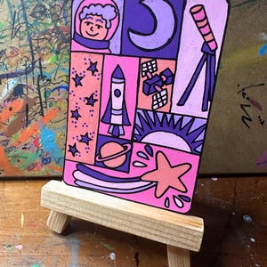 May include: A colorful space-themed painting on a small wooden easel. The artwork features an astronaut, moon, stars, rocket, planet, satellite, sun, and shooting star in shades of pink, purple, and orange. The painting is rectangular and has a black border.