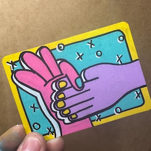 May include: A colourful illustration of two hands clasped together on a rectangular card. One hand is pink with yellow fingernails, and the other is purple. The background is teal with yellow borders and white accents.