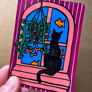 Puede incluir: Ilustración pintada a mano de un gato negro sentado en una ventana, mirando un pez dorado. La ventana es rosa, con un cielo azul. Una planta verde cuelga de la ventana. El fondo es de rayas magenta.