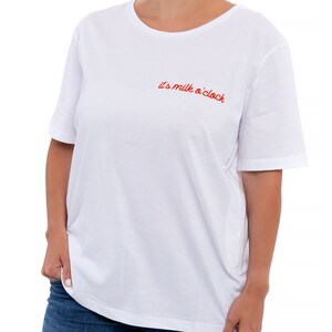 Puede incluir: Camiseta blanca de manga corta y cuello redondo. La camiseta tiene un texto bordado en rojo que dice "it's milk o'clock".
