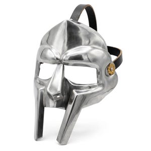 May include: A polished silver-colored metal face mask featuring a prominent central nose guard, wide eye openings, and two vertical chin bars. The mask is secured with a black leather strap attached to the sides by small, decorative gold-toned floral rivets.