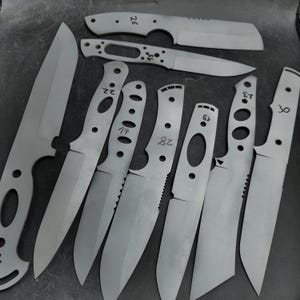 Op de afbeelding: Een assortiment onafgewerkte metalen mesbladen, in verschillende vormen en maten, gerangschikt op een donker oppervlak. Sommige bladen hebben gaten in de handvatten en andere hebben gekartelde randen. Er staan nummers op sommige bladen.