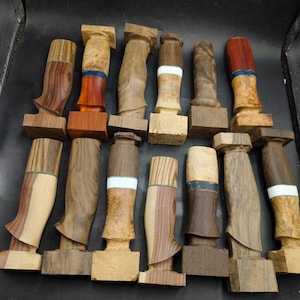 Puede incluir: Una colección de mangos de cuchillos de madera en varias formas y vetas. Algunos mangos presentan bandas de color contrastantes, incluyendo rojo, azul y blanco. Los mangos se exhiben sobre una superficie oscura, mostrando sus diseños únicos.