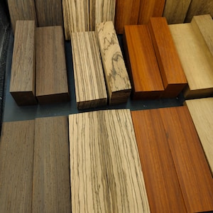 May include: Assortment of wood blanks in various colors and grain patterns. The blanks are rectangular and have a smooth finish. They are arranged in rows on a black surface.