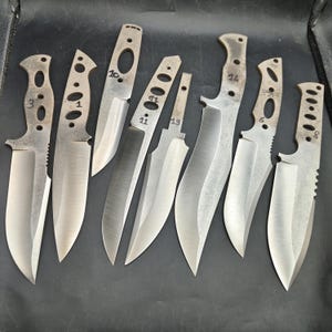 May include: A collection of eight unfinished knife blades, each with a silver, metallic finish. The blades vary in shape and size, with some featuring holes and textured handles. Numbers are etched on some of the blades.