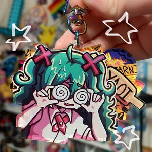 May include: A colorful anime-style keychain featuring a character with teal hair, spiral eyes, and a pink outfit. The keychain has a rainbow-colored clasp and includes star-shaped and text-based accents.