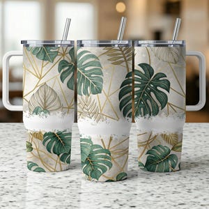 Op de afbeelding: Drie roestvrijstalen reisbekers met handvatten en rietjes. De bekers hebben een witte band en zijn versierd met groene monstera bladeren, gouden geometrische lijnen en beige bladeren tegen een beige achtergrond.