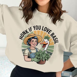 Puede incluir: Sudadera color crema con un gráfico circular que presenta a una mujer con sombrero de paja y peto, sosteniendo una planta de albahaca en maceta junto a un ganso sonriente. El texto "HONK IF YOU LOVE BASIL" está arqueado sobre la imagen.