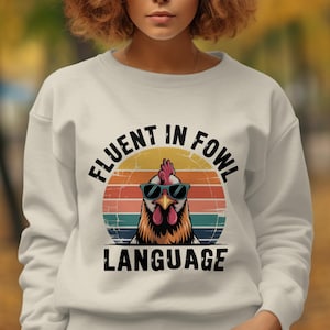 Pode incluir: Camisola branca com um desenho gráfico de um galo com óculos de sol, contra um fundo de pôr do sol. O texto "FLUENT IN FOWL LANGUAGE" está arqueado acima e abaixo da imagem do galo. Um estilo de camisola casual e confortável.