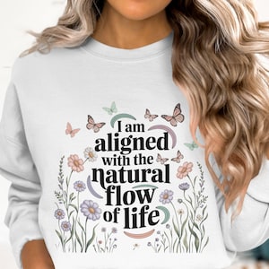 Peut inclure: Sweat-shirt blanc avec l'inscription « I am aligned with the natural flow of life ». Le motif comprend des papillons, des fleurs et des éléments botaniques aux couleurs pastel. Un haut décontracté et confortable.
