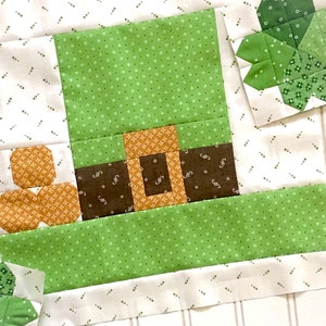 May include: A green, brown, and orange patchwork quilt block featuring a leprechaun's hat with a buckle and a shamrock. The block is surrounded by a white background with a small brown pattern.