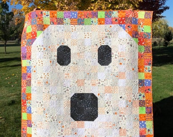 Puede incluir: Una colcha blanca de patchwork con un diseño de fantasma. El fantasma tiene ojos negros y una boca. La colcha está rodeada por un borde de patchwork colorido.