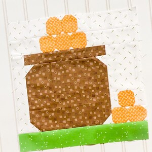 May include: A brown pot with a white and brown patterned design, filled with orange and white patterned circles. The pot is sitting on a green strip with white polka dots.