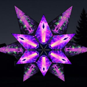 Puede incluir: Una gran decoración en forma de estrella iluminada con un diseño complejo y multifacético. La estrella es principalmente púrpura con acentos de rosa y verde, y presenta un núcleo central de múltiples puntas. El fondo es un cielo oscuro y nublado con siluetas de árboles y una casa.