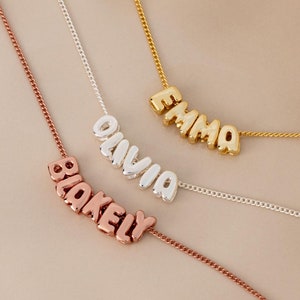 Könnte beinhalten: Drei Halsketten in Gold, Silber und Roségold mit den personalisierten Namen "Emma", "Olivia" und "Blakely", die in Blasenschrift geschrieben sind.
