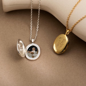 Pode incluir: Dois colares com pingentes. Um é prateado, redondo e aberto, revelando uma pequena foto. O outro é dourado, oval e fechado, com um design floral. Ambos pendem de correntes delicadas, mostrando um estilo de joalheria clássico.