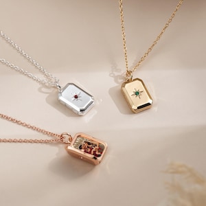 Könnte beinhalten: Drei rechteckige Medaillon-Halsketten an feinen Ketten. Ein silbernes Medaillon mit rotem Sternstein, ein goldenes Medaillon mit grünem Sternstein und ein offenes roségoldenes Medaillon, das ein kleines Foto von zwei Personen im Inneren zeigt.